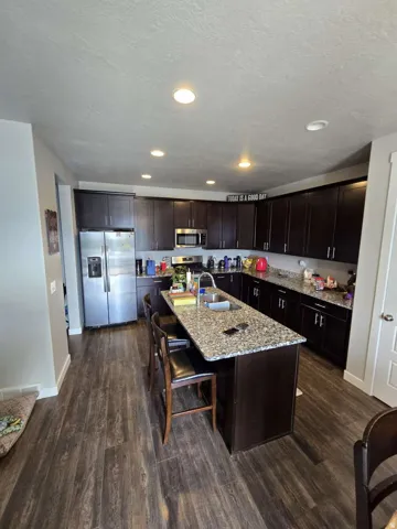 Kitchen featuring dark wood-finish cabinetry, granite countertops, stainless steel appliances, a center island with integrated sink, and wood-finish flooring