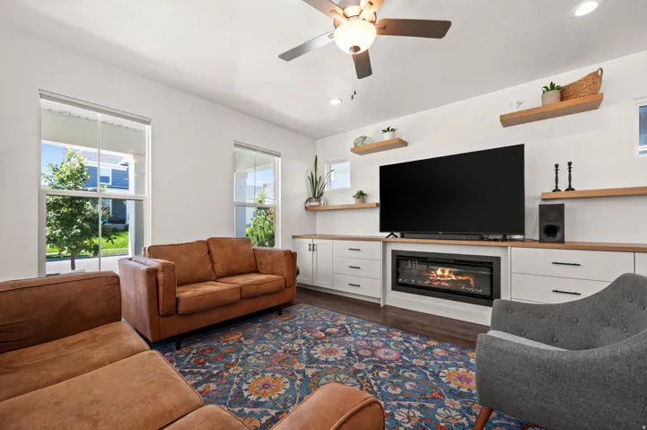Built-in media console with fireplace, wood-finish flooring, two large windows, white walls, and a ceiling fan