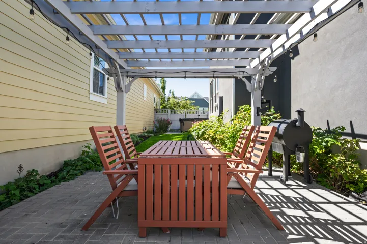 Paved patio featuring a white pergola, string lighting, and an adjacent green space
