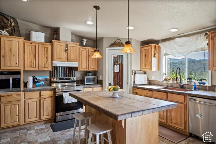 Wood-finish cabinetry, stainless steel appliances, central island with tiled countertop, recessed lighting, and expansive window with outdoor scenery