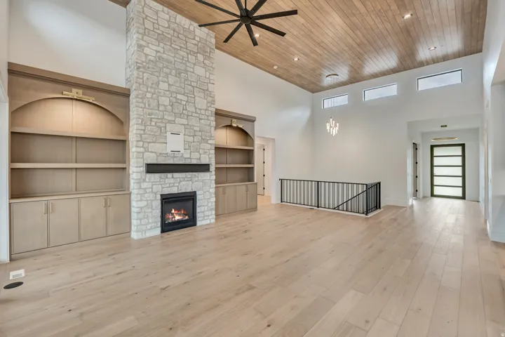 Vaulted ceiling with wood-finish planks, stone fireplace, built-in cabinetry with arched shelving, light wood-finish flooring, and black metal railing