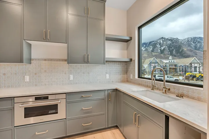 Expansive window framing a mountain backdrop, gray shaker-style cabinetry with gold-finish hardware, built-in stainless steel microwave, large single-basin sink with gooseneck faucet, and light-toned backsplash