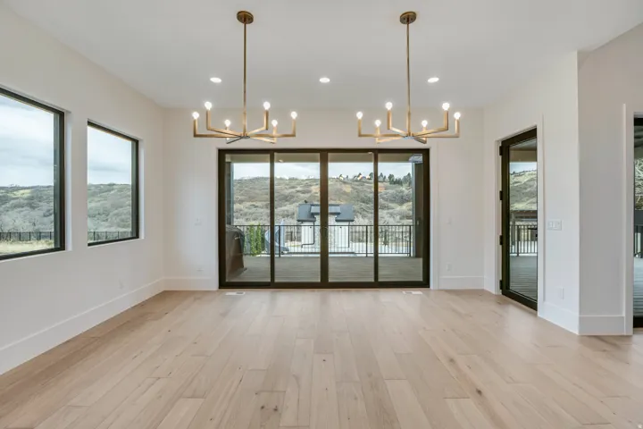 Spacious room with wood-finish flooring, two modern chandeliers, multiple windows, and a glass-paneled sliding door leading to an outdoor balcony