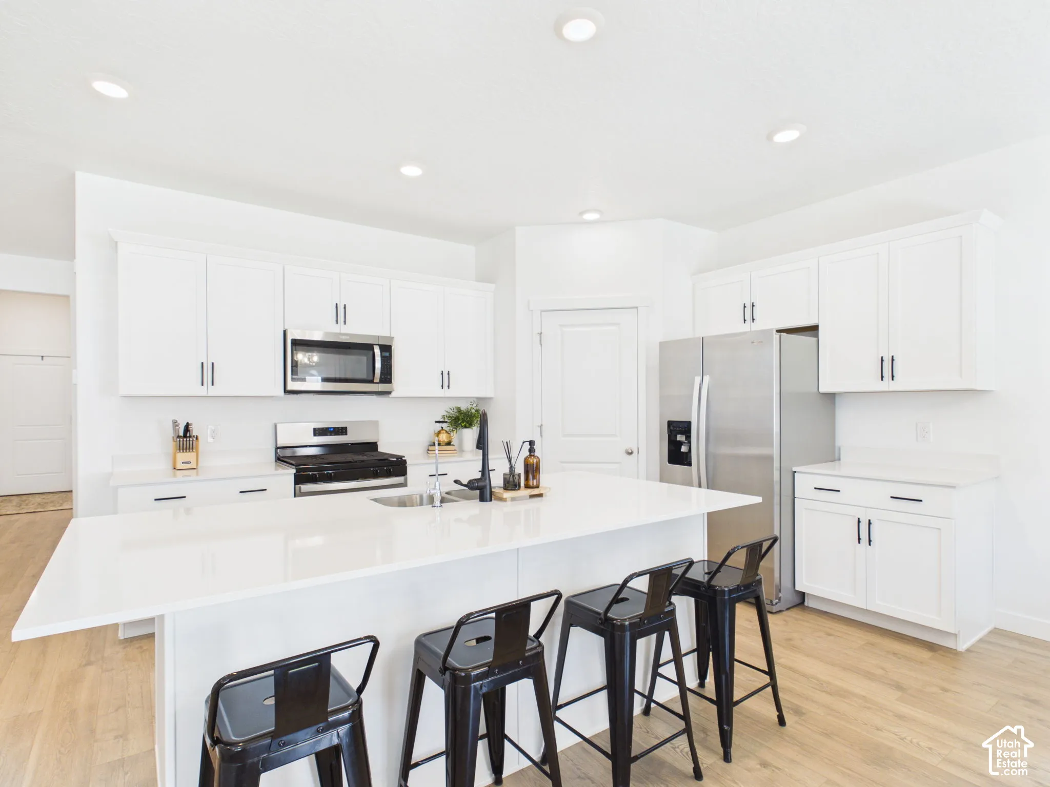 White cabinetry with matte black hardware, stainless steel appliances, a center island with counter seating, light wood-finish flooring, and recessed ceiling lights