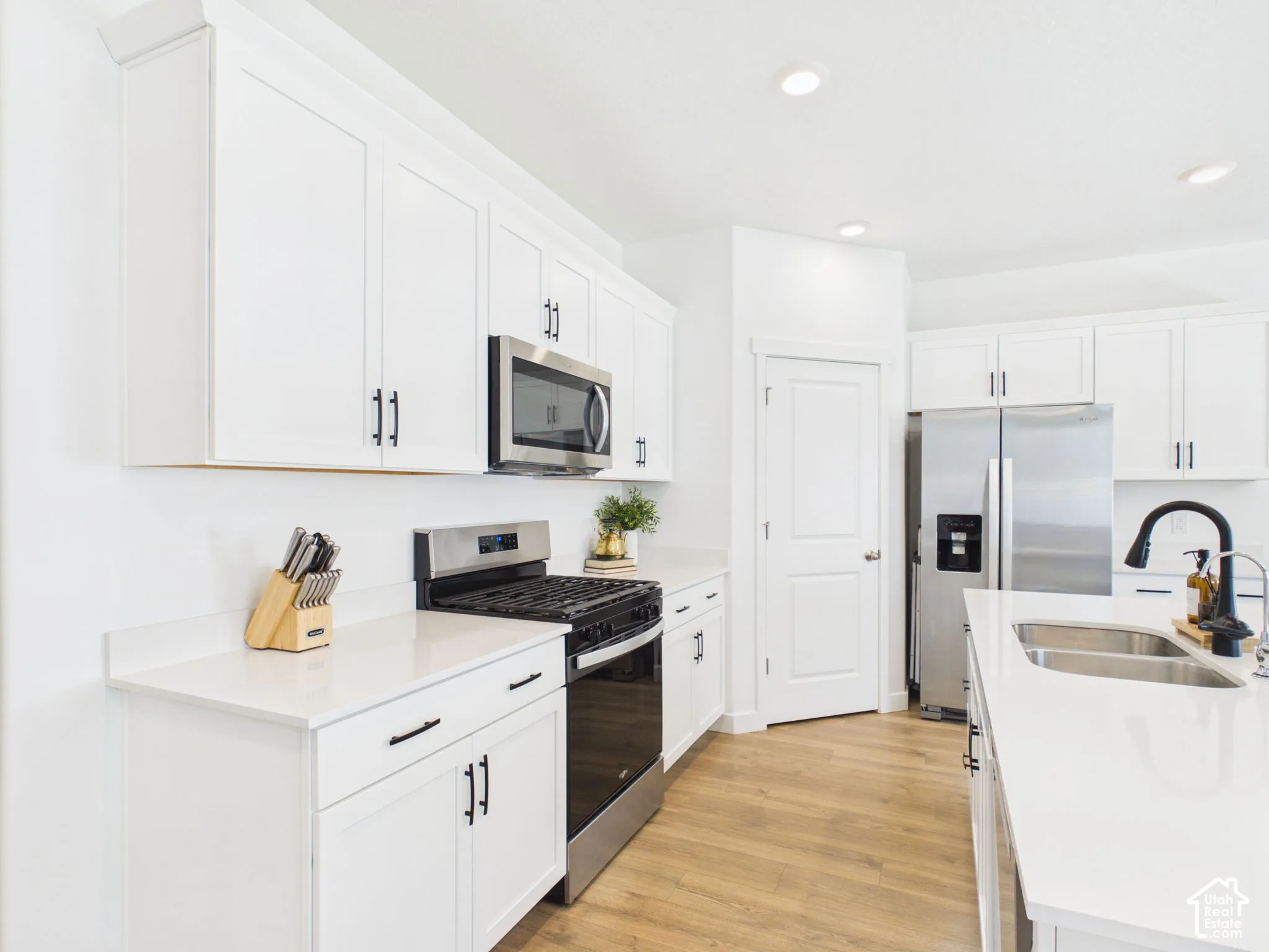 White shaker cabinetry with black hardware, stainless steel appliances, light-toned wood-finish flooring, recessed lighting, and white countertops