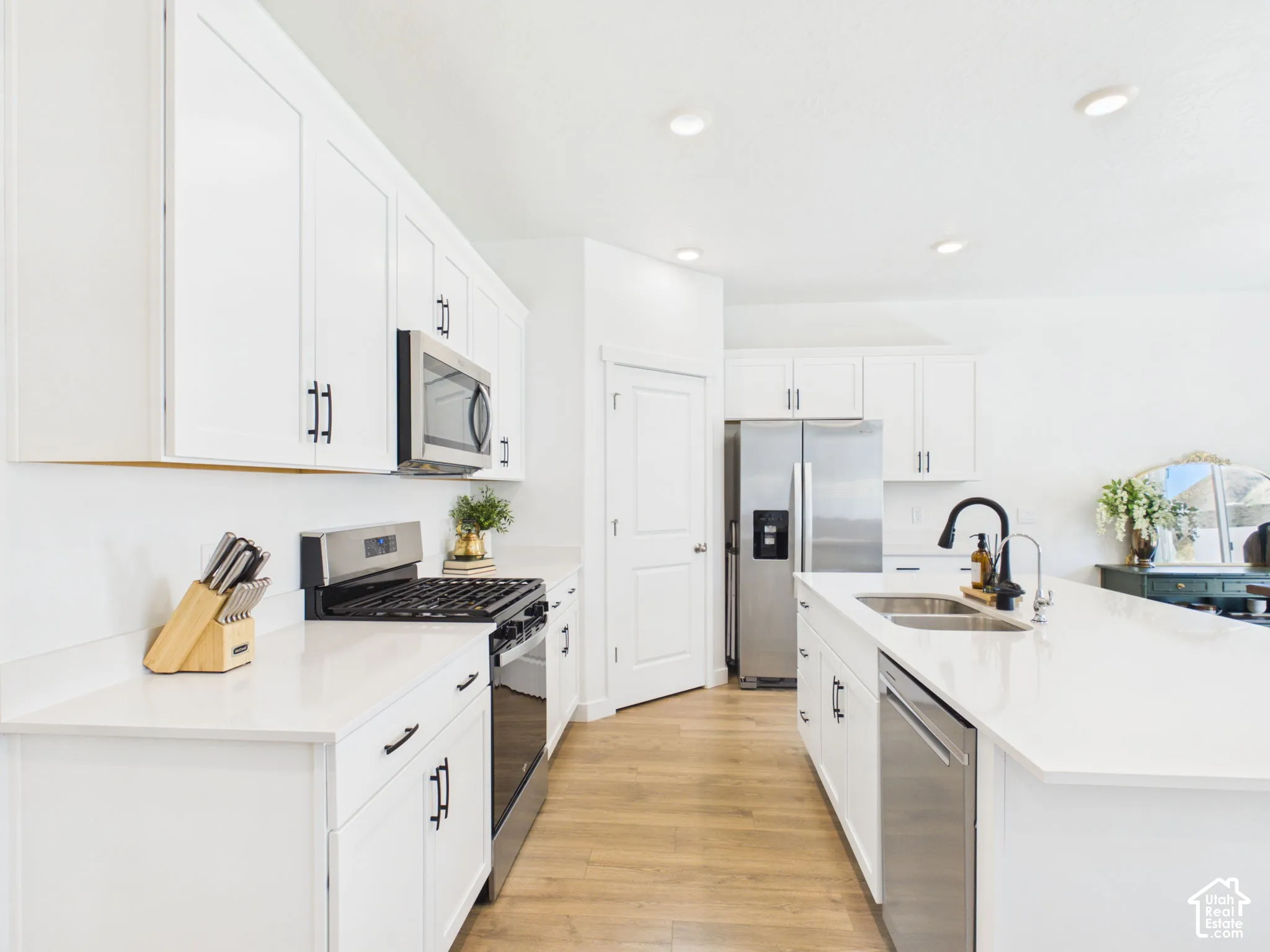 White cabinetry with matte black hardware, stainless steel appliances, light-toned wood-finish flooring, white countertops, and recessed lighting