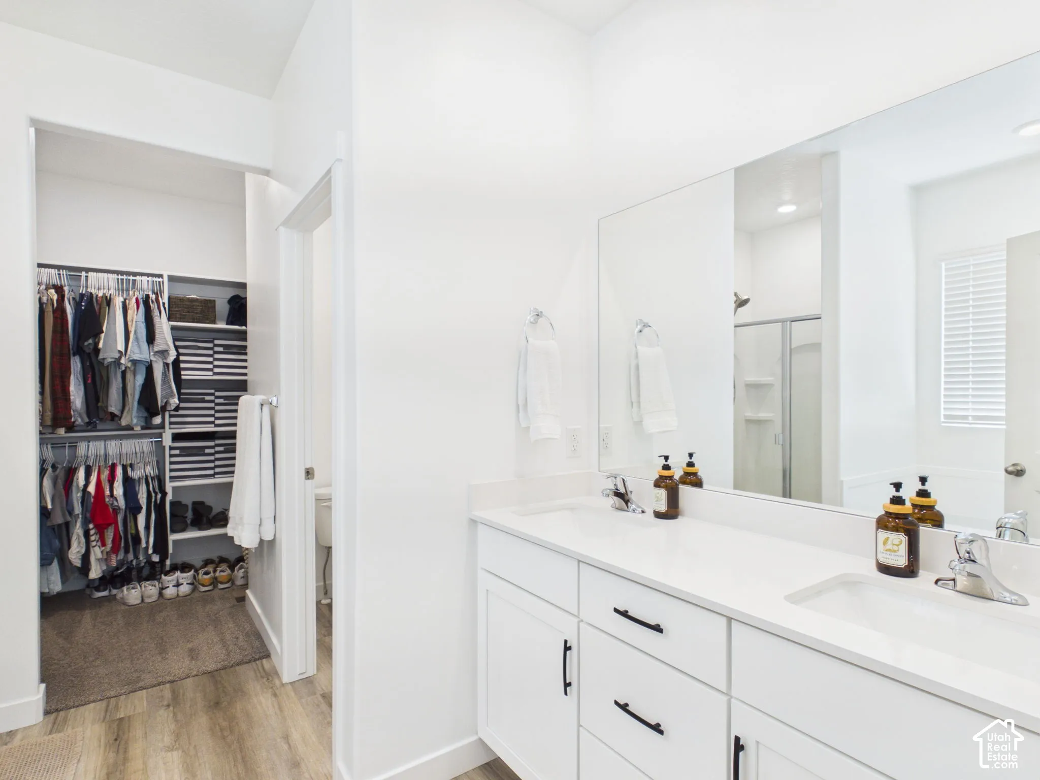 Dual vanity with white cabinetry and black hardware, a large wall-mounted mirror, and wood-finish flooring