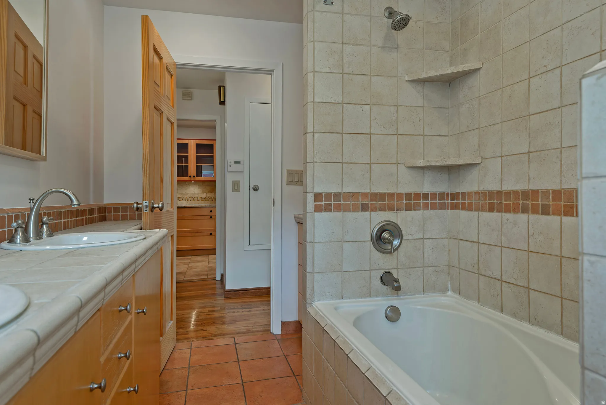 Bathroom featuring a built-in bathtub, a shower with integrated shelving, tile flooring, a wood-finish vanity with dual sinks, and a tile countertop
