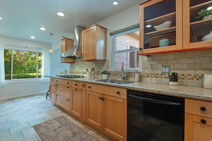 Kitchen featuring natural wood cabinetry, light-toned countertops, a stainless steel range hood, a tile backsplash with a mosaic accent, and recessed lighting