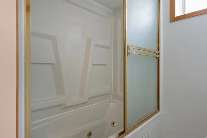 White tub with integrated shower surround, brass-finish framed frosted glass shower doors, and overhead window