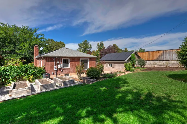Expansive lawn area with a brick residence, a detached structure featuring solar panels, and multiple raised garden beds