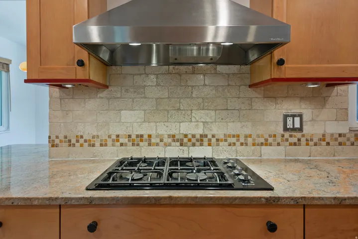 Built-in stainless steel range hood, stone tile backsplash with decorative mosaic accent, cooktop, wood cabinetry, and granite countertops