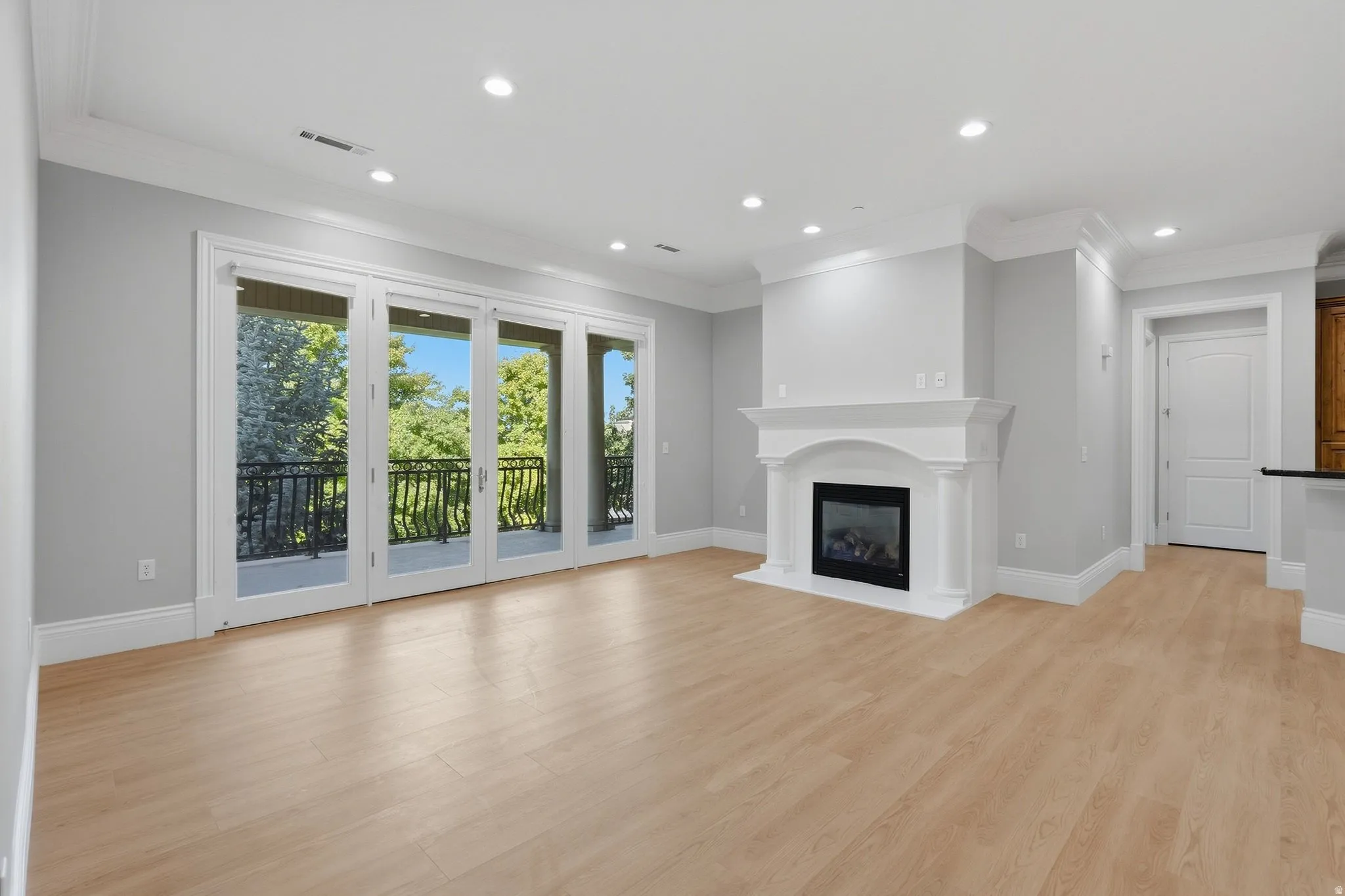 Expansive living area with light wood-finish flooring, a classic fireplace with a decorative mantel, crown molding, recessed lighting, and a private balcony with wrought iron railings