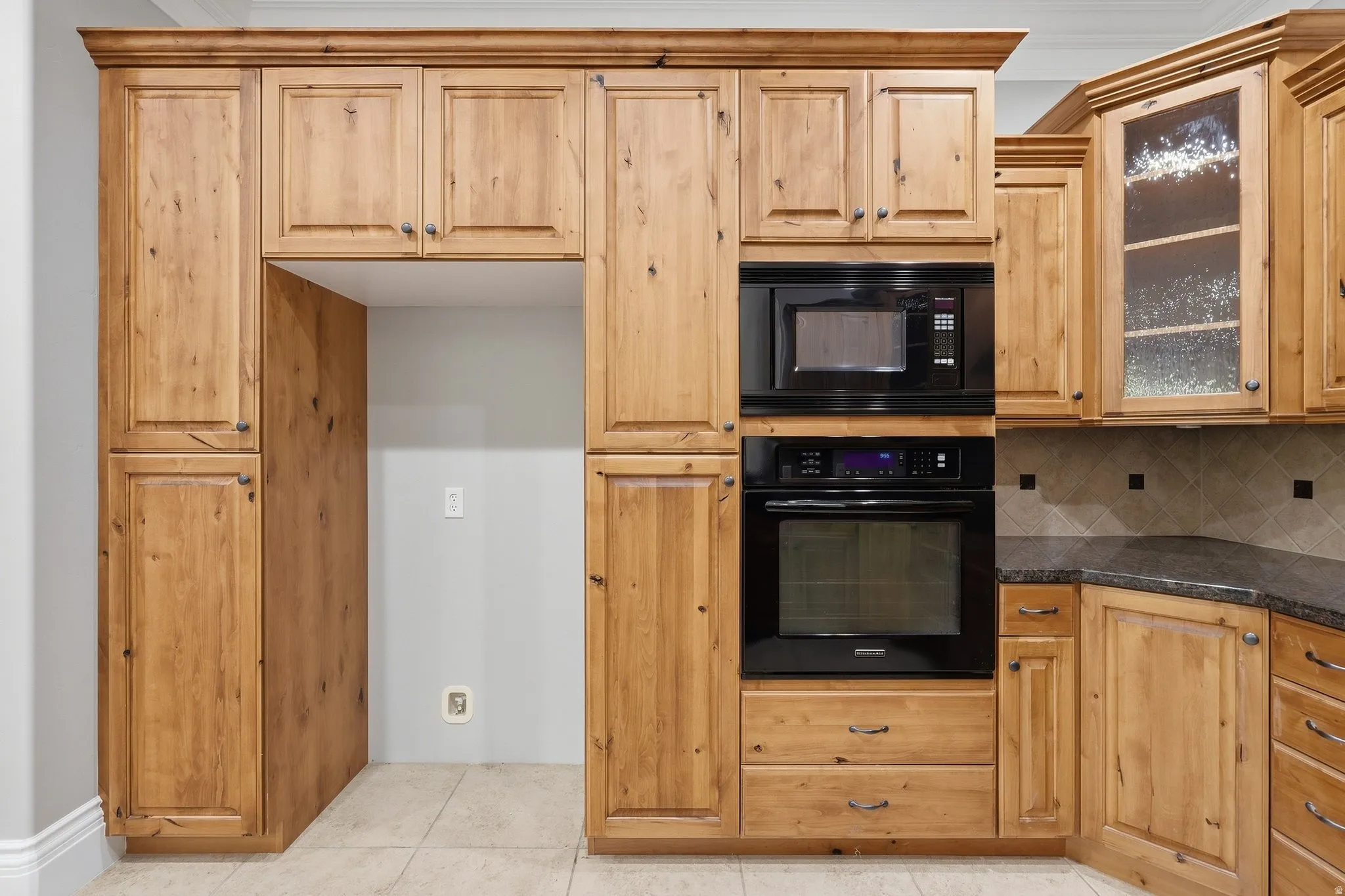 Built-in oven and microwave, natural wood cabinetry, glass-front upper cabinets, tile flooring, and dark stone countertops
