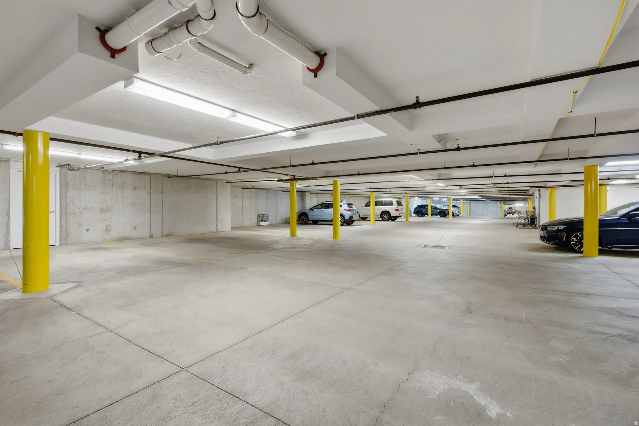 Underground parking garage with concrete flooring, exposed ceiling piping, and fluorescent lighting
