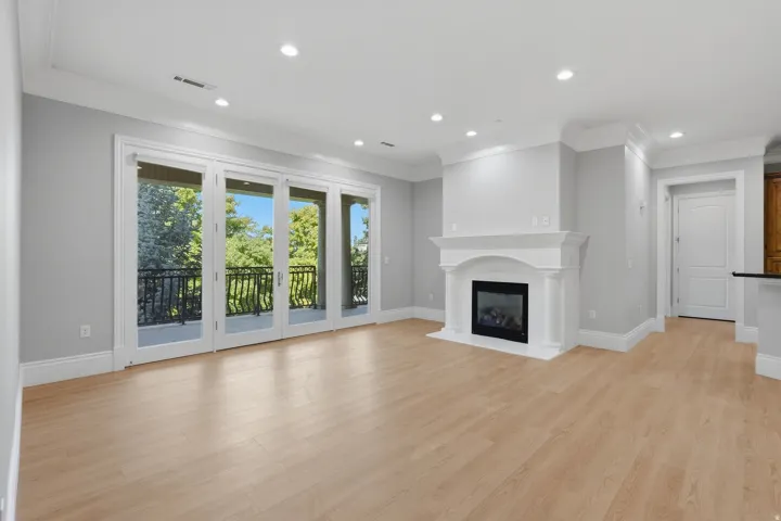 Expansive living area with light wood-finish flooring, a classic fireplace with a decorative mantel, crown molding, recessed lighting, and a private balcony with wrought iron railings