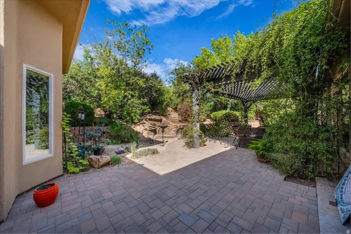 Paver patio with a covered pergola featuring an established vine, built-in rock landscaping, and mature trees