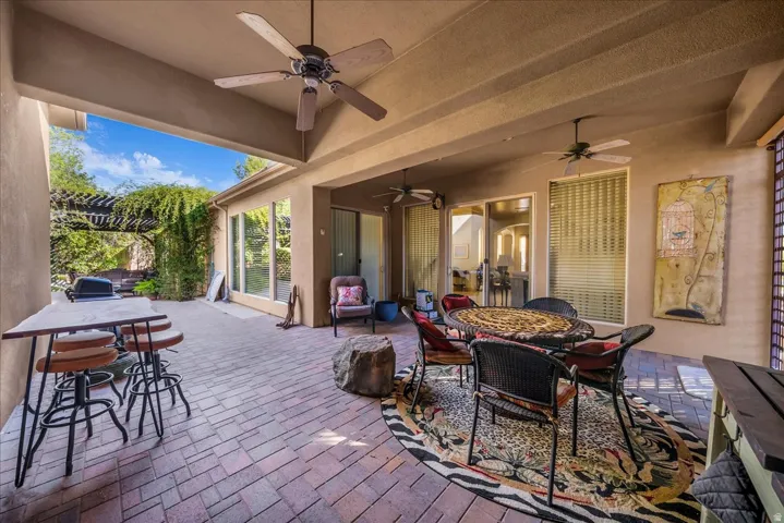 Expansive covered patio featuring brick-patterned hardscaping, multiple ceiling fans, full-height glass doors, and an attached wood pergola with mature climbing foliage