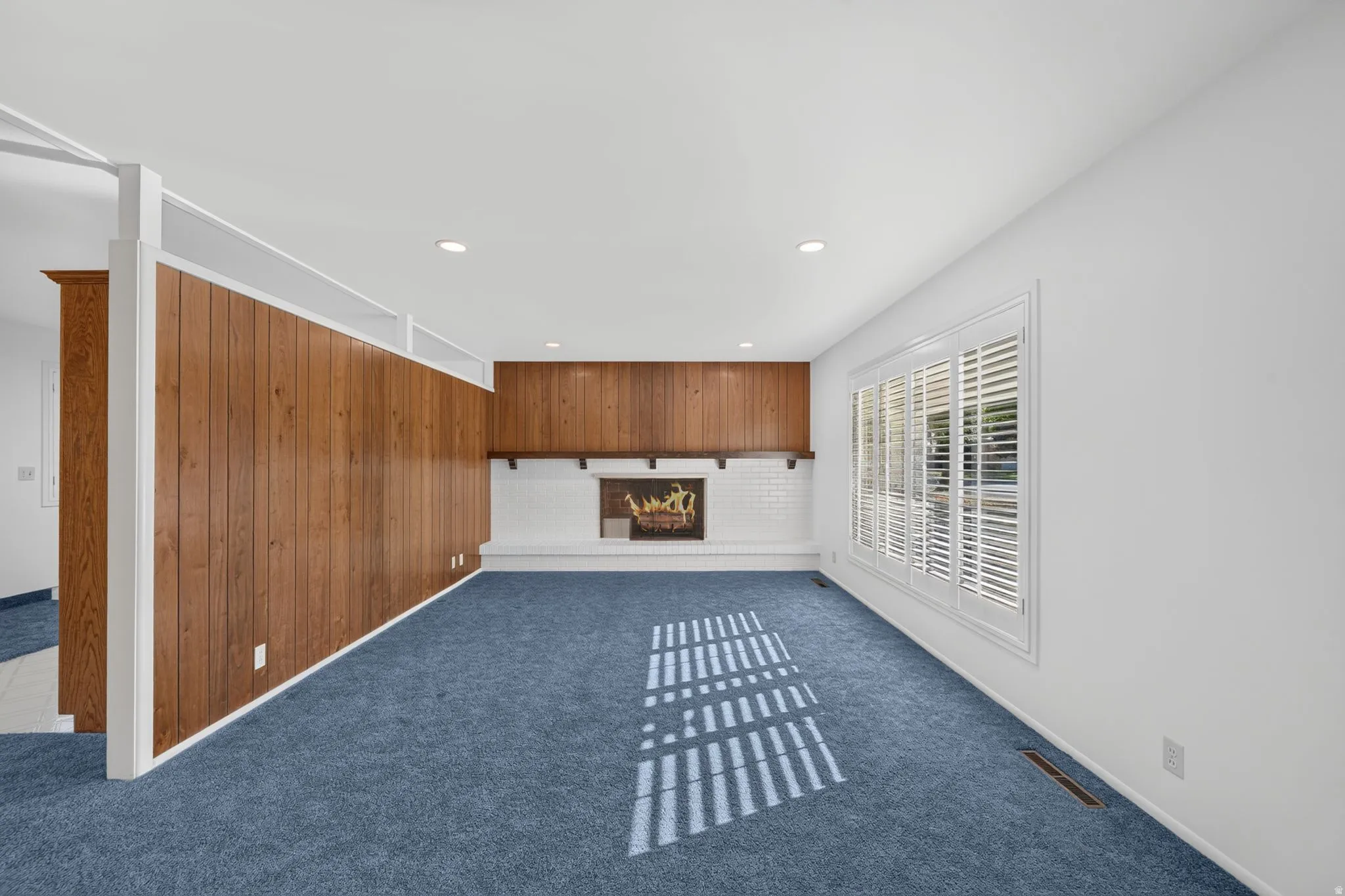 Recessed lighting, white walls, wood-paneled accent wall, white brick fireplace with mantel, and a window with white plantation shutters