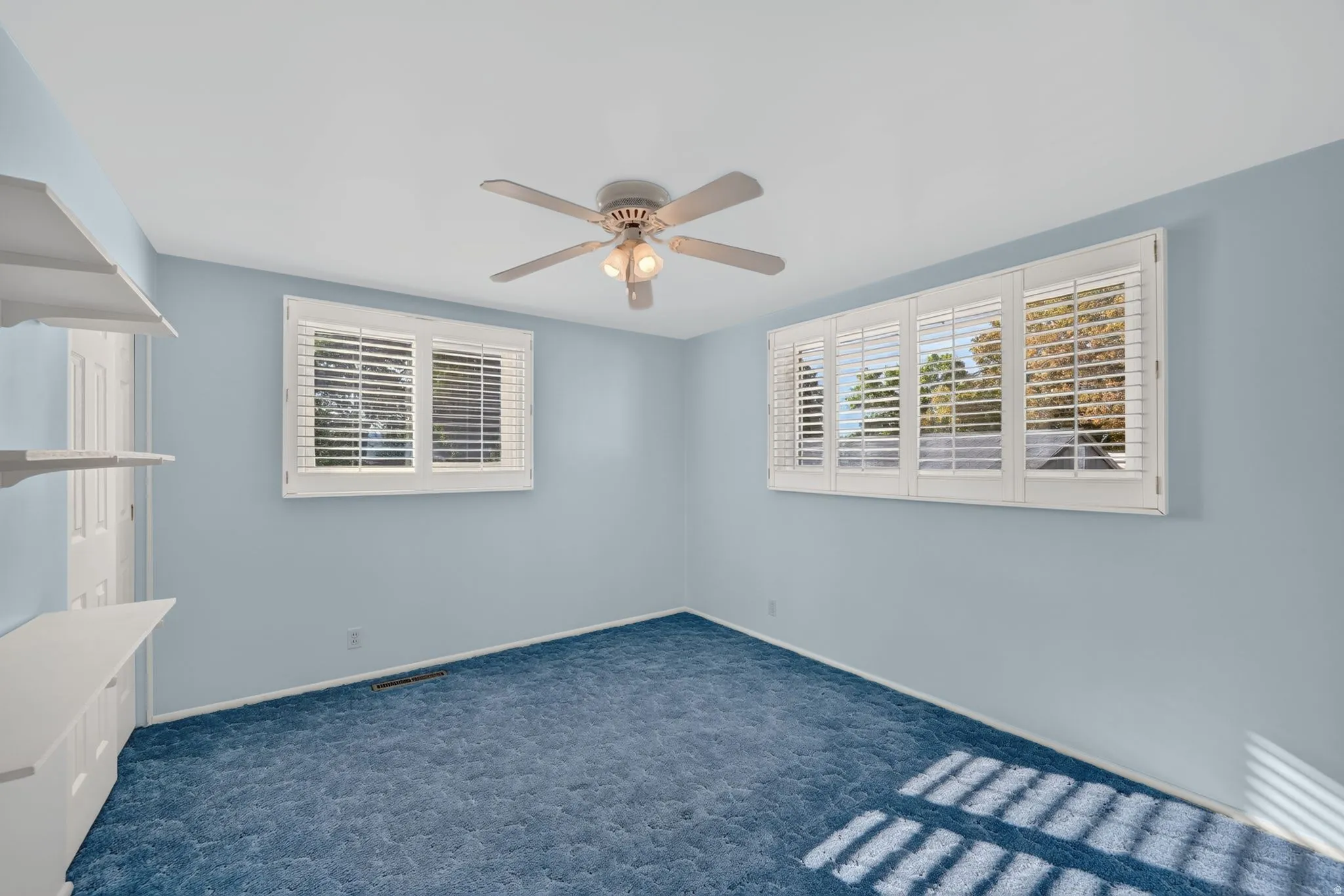 Expansive wall with white plantation shutters, two windows, and a ceiling fan with integrated lighting