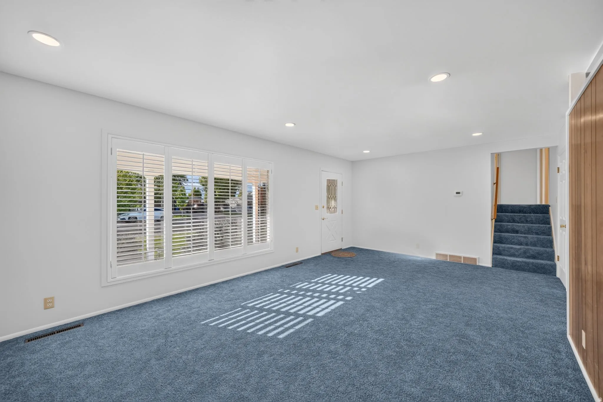 Recessed lighting, white walls, blue carpeting, white plantation shutters, and a wood-paneled accent wall