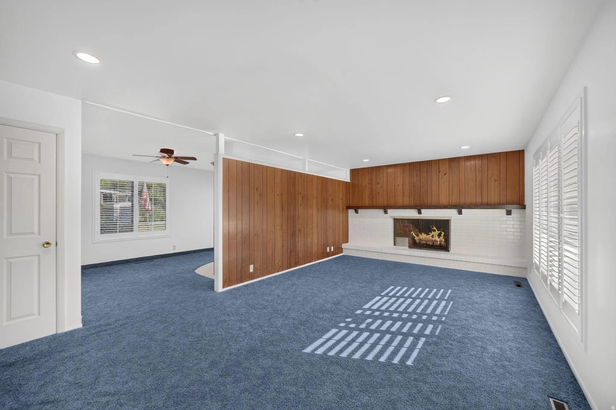 Room featuring a white brick fireplace with a wood mantel, wood paneling, recessed lighting, and white plantation shutters