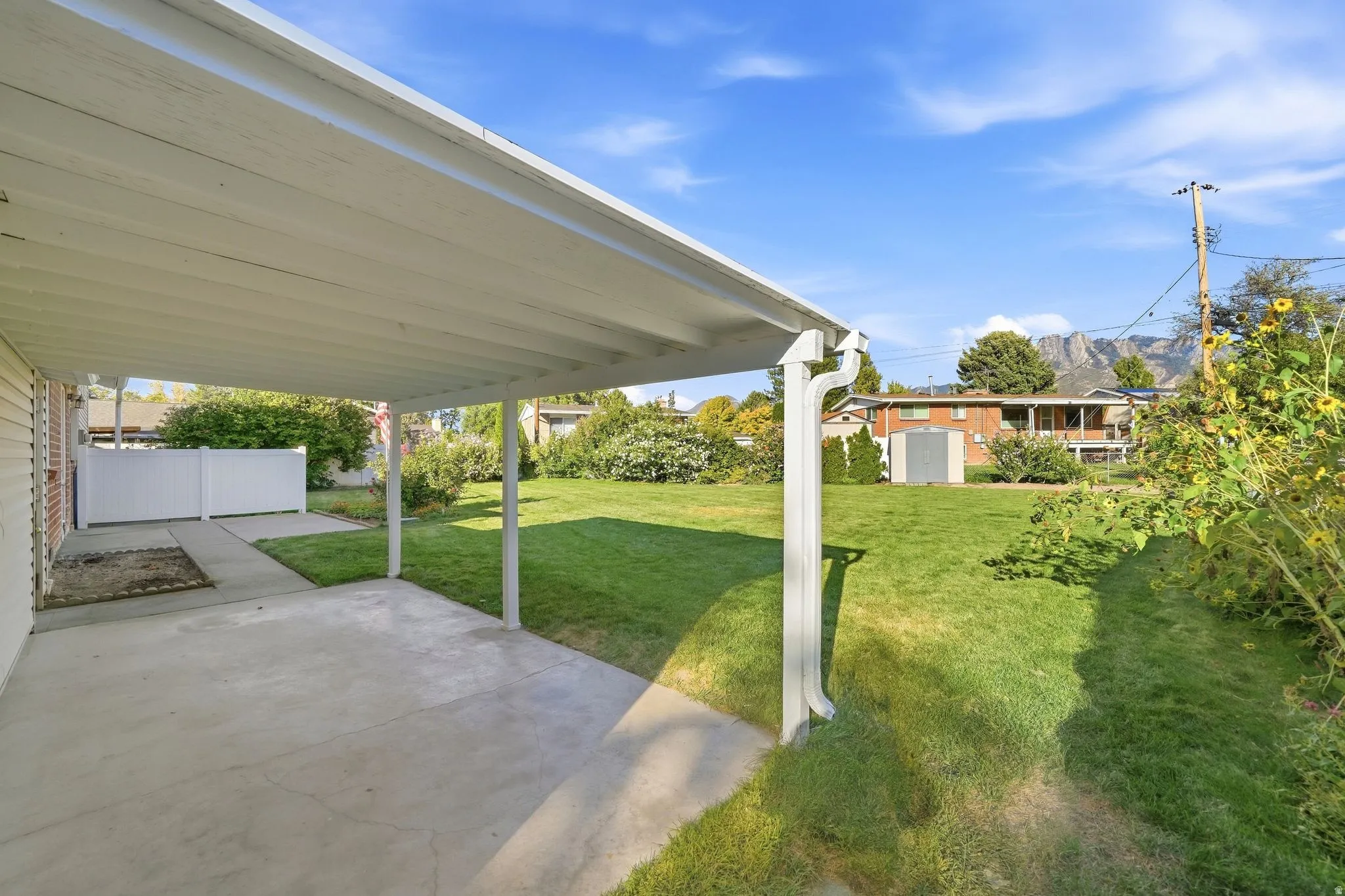 Covered concrete patio with a spacious lawn, white privacy fencing, and a storage shed