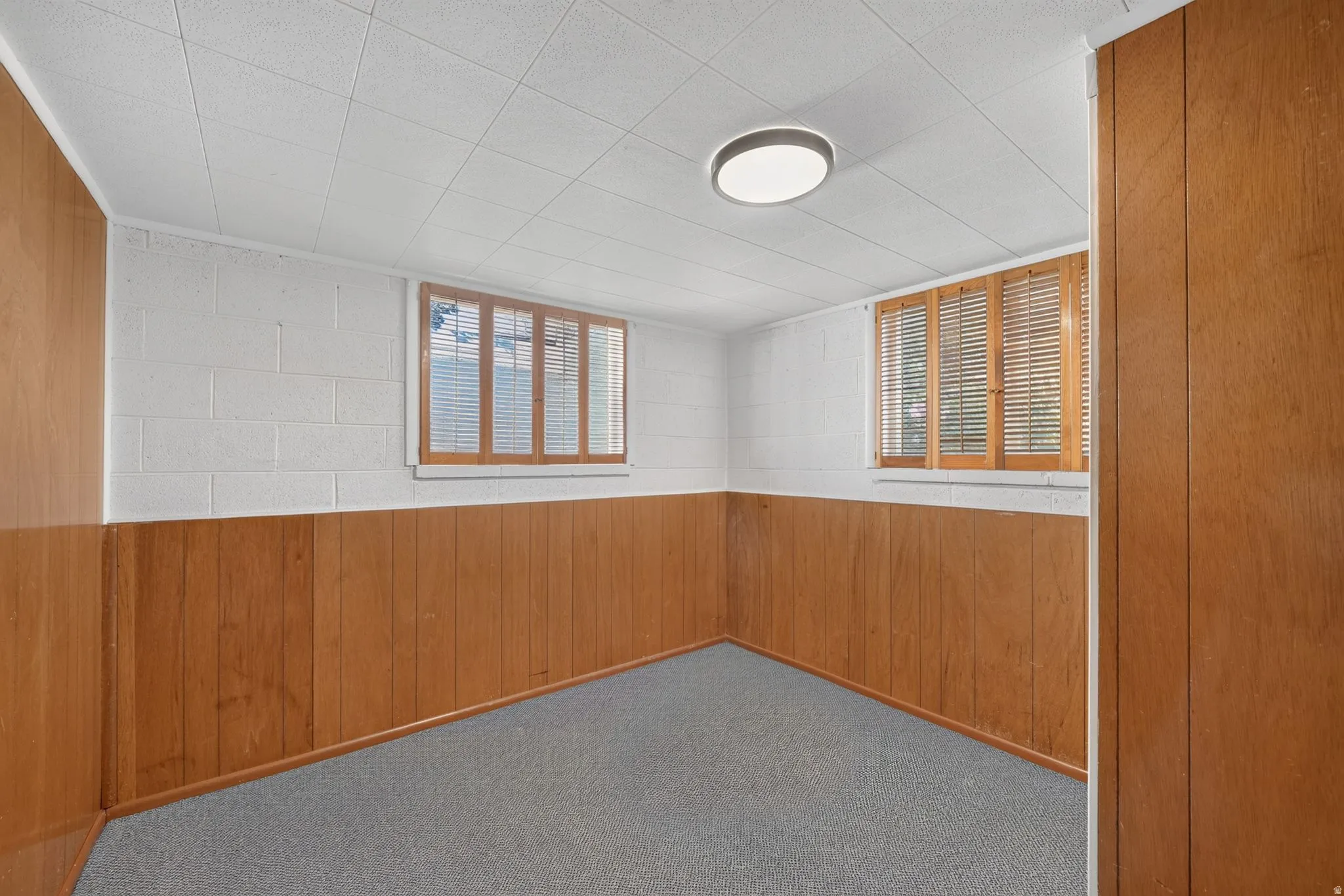 Recessed lighting, wood-finish paneling, two windows with wood blinds, white block walls, and gray carpet flooring