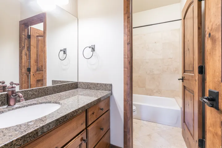 Granite countertop with an undermount sink, rustic wood-finish cabinetry, built-in vanity mirror, brushed bronze-finish faucet, and a tiled shower surround