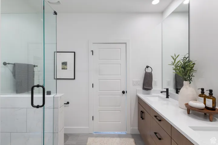 Walk-in shower with clear glass enclosure and tiled surround, dual vanity with wood-finish cabinetry, white countertop, and matte black fixtures, full-height wall mirror, and recessed lighting