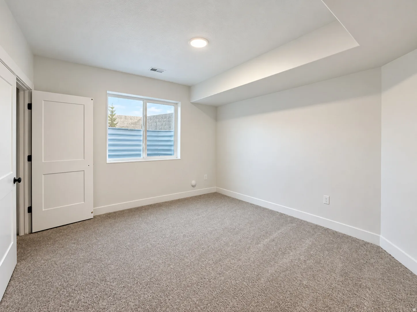 Light-colored carpeting, white walls, and a window with a corrugated metal window well