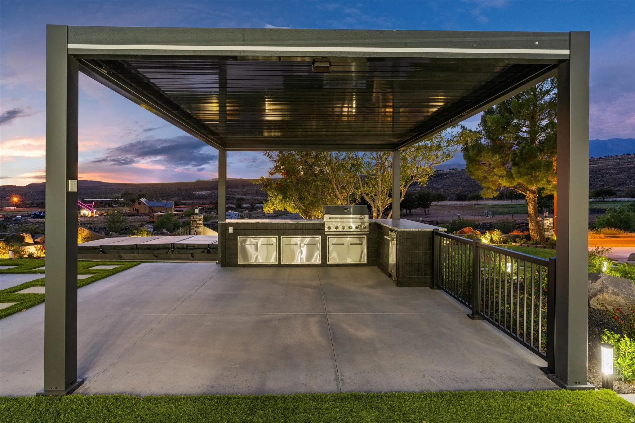 Contemporary outdoor kitchen featuring an integrated grill and stainless steel cabinetry, set under a louvered pergola with recessed lighting, on a concrete patio with a black metal railing
