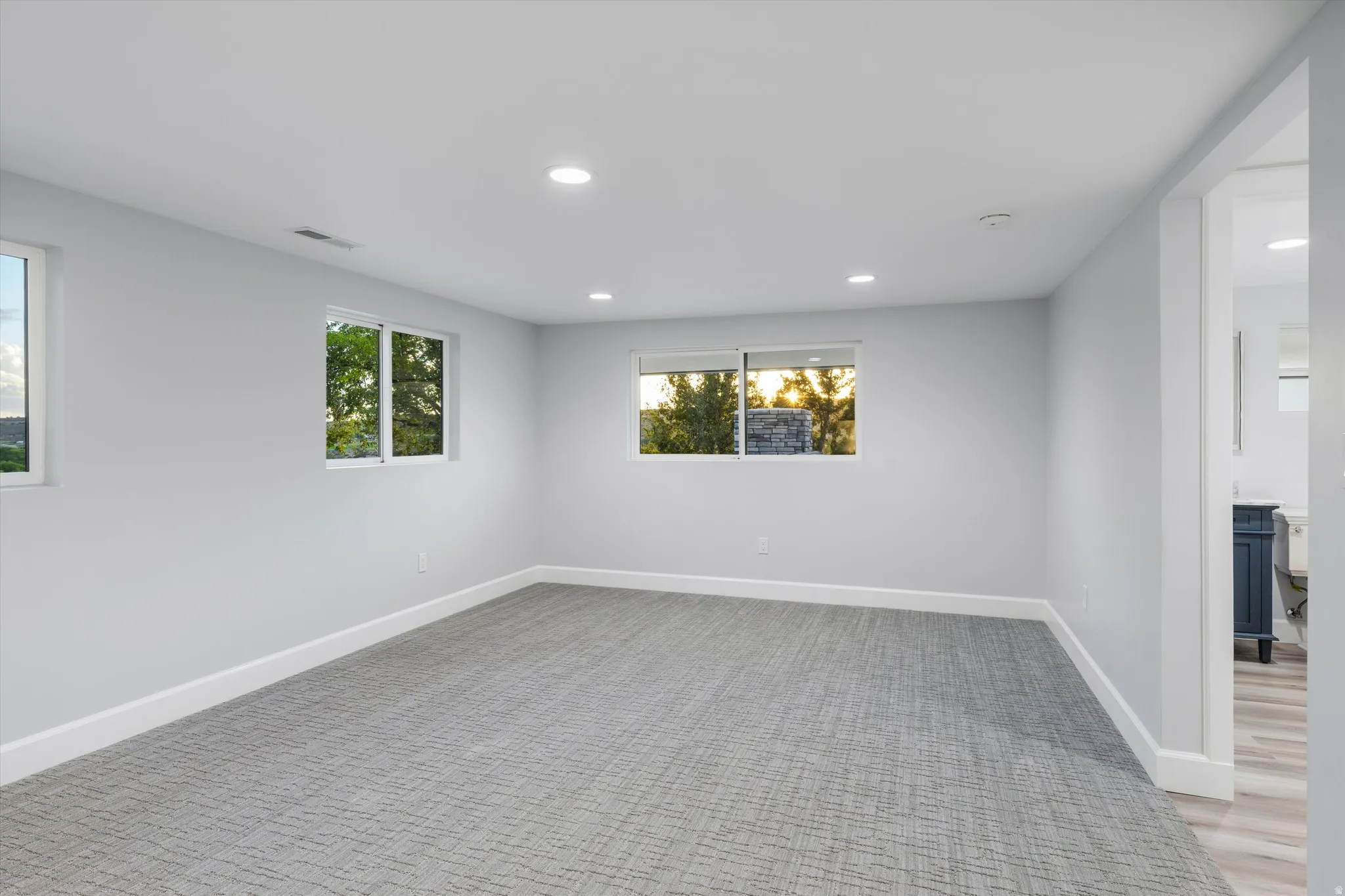 Recessed lighting, light-colored walls, multiple windows, patterned carpeting, and an adjoining space with wood-finish flooring