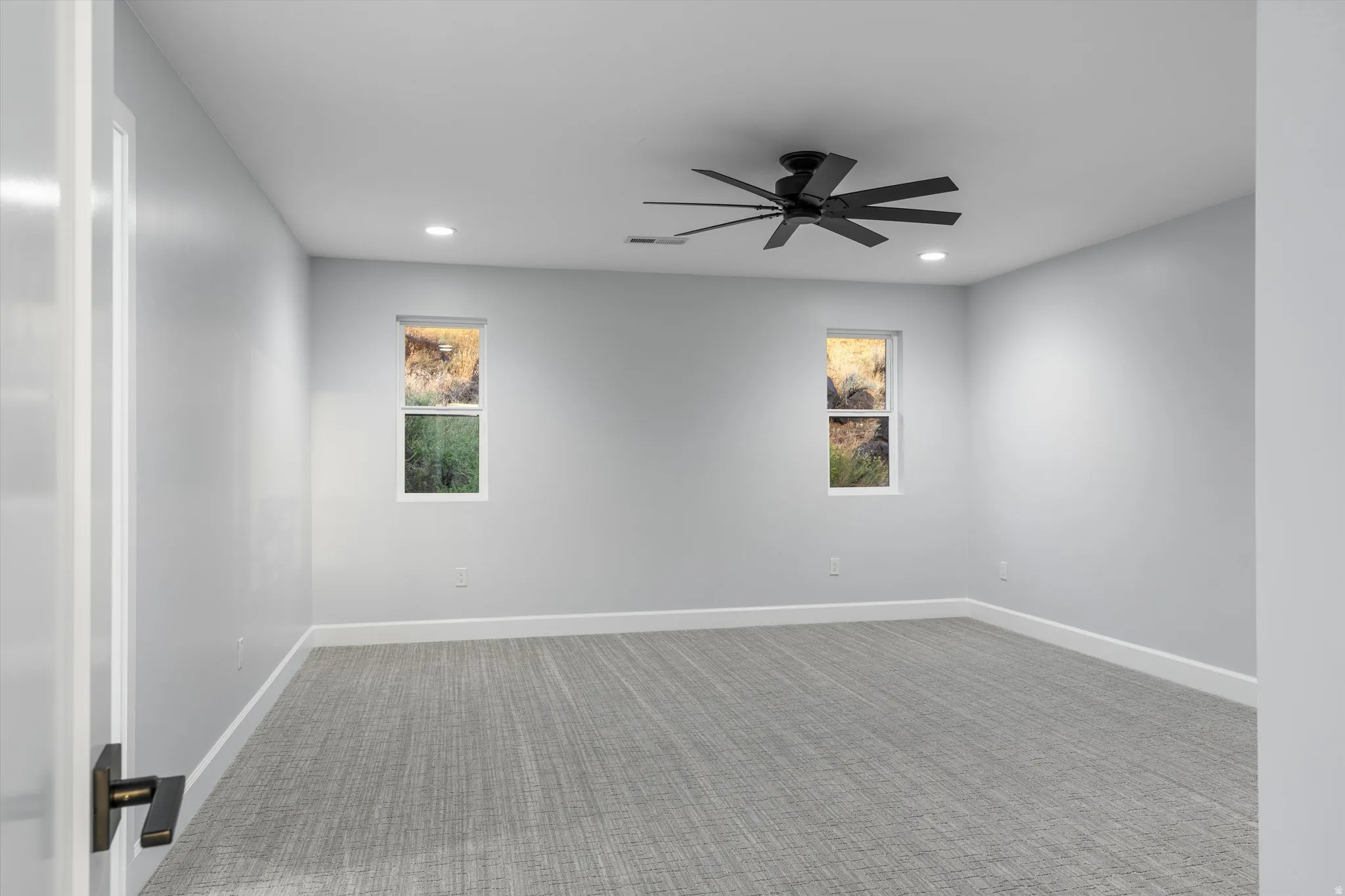 Recessed lighting, ceiling fan with matte black blades, two rectangular windows, gray patterned carpeting, and white trim
