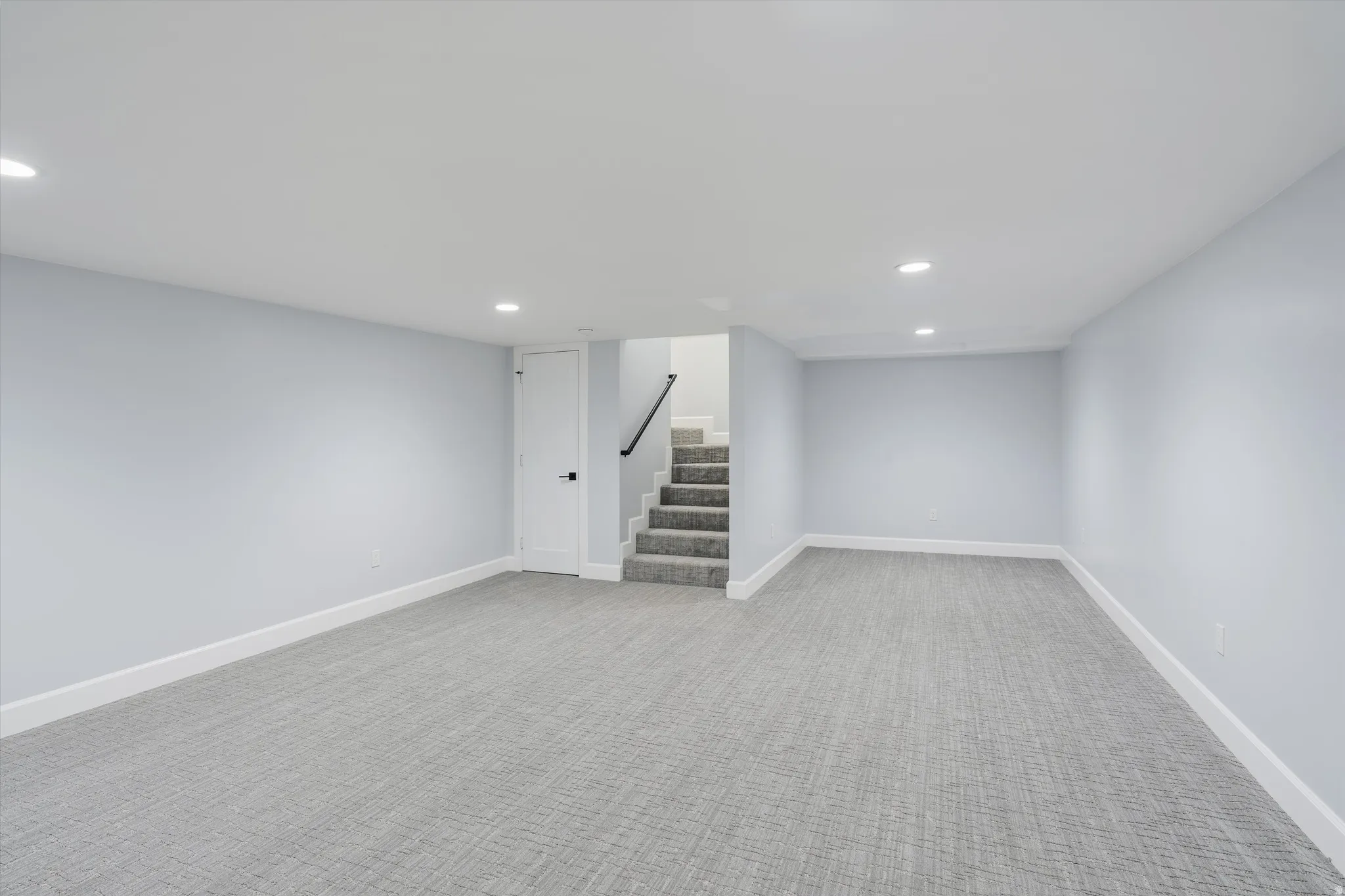 Spacious room with recessed lighting, light gray walls, gray patterned carpeting, white trim, and a staircase with a black handrail