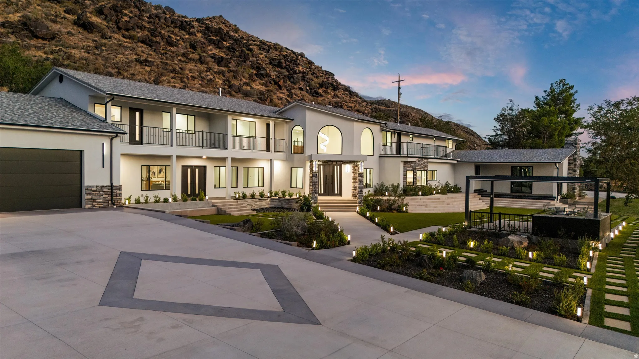 Gated driveway with geometric design, multi-level exterior with stone accents, integrated lighting, upper-level balconies, and a structured landscaping design
