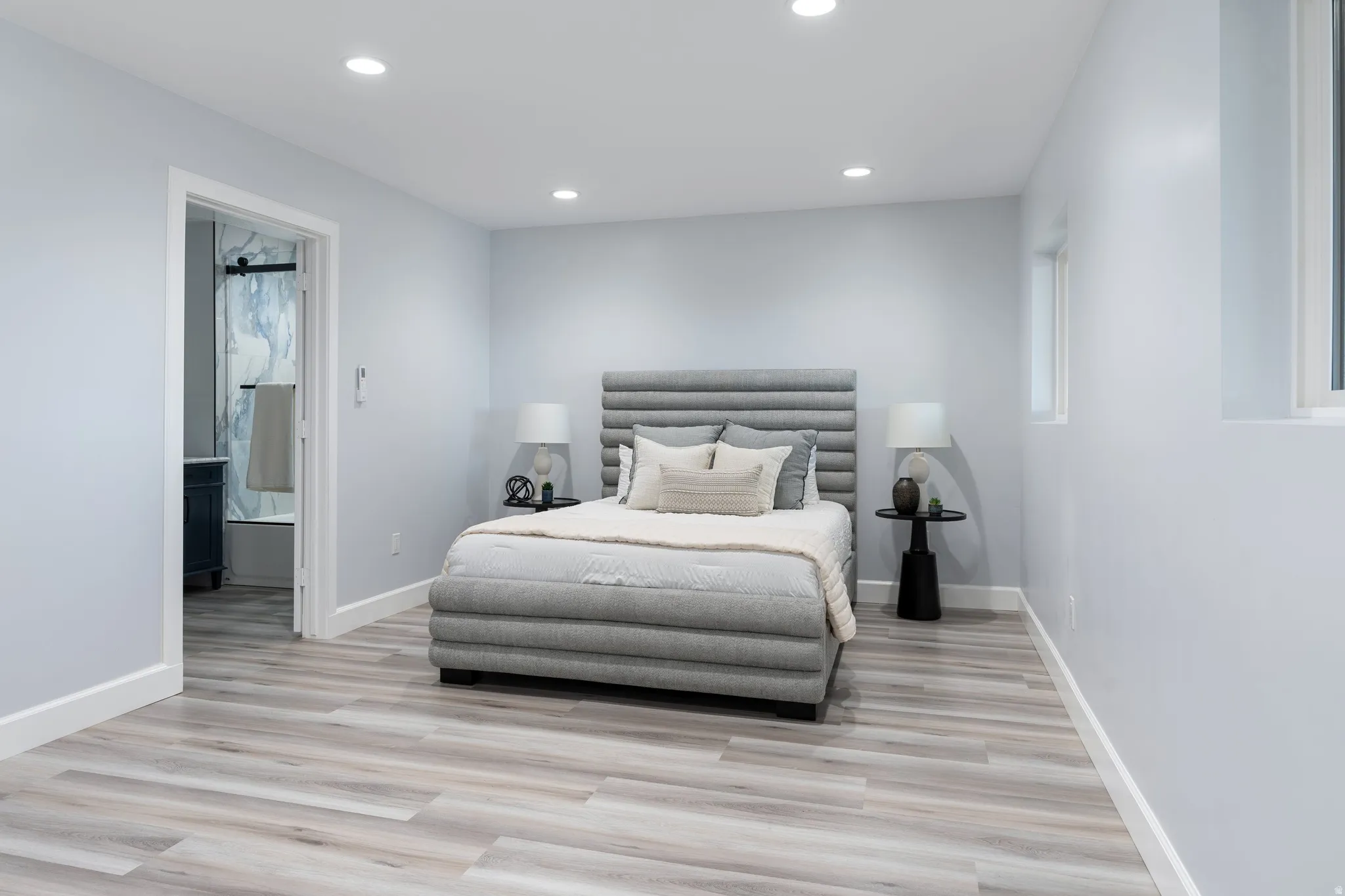 Wood-finish flooring throughout, recessed ceiling lighting, light-toned wall paint, and a doorway to a full bathroom with a dark wood-finish vanity