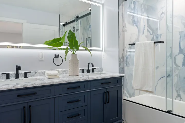 Dual vanity with granite countertop, matte black fixtures, integrated lighted mirror, and deep blue cabinetry