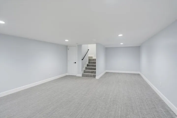 Spacious room with recessed lighting, light gray walls, gray patterned carpeting, white trim, and a staircase with a black handrail