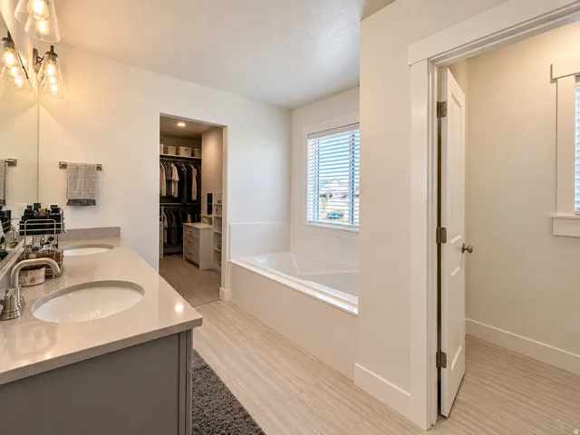 Dual vanity with integrated sinks and brushed nickel faucets, deep soaking tub with a window, tile flooring, and a walk-in closet with built-in shelving