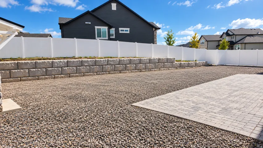 Gravel backyard with a paver patio area, retaining wall, and white privacy fencing