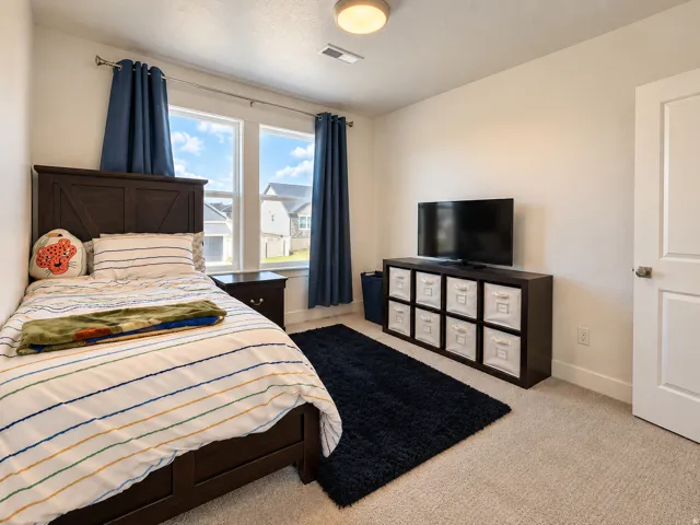 Recessed ceiling light, neutral carpet flooring, two pane windows, wood-finish headboard, and a white interior door with a brushed nickel handle