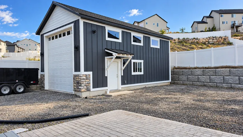 Detached structure with white panel garage door, dark vertical siding, white trim, and stone veneer accents