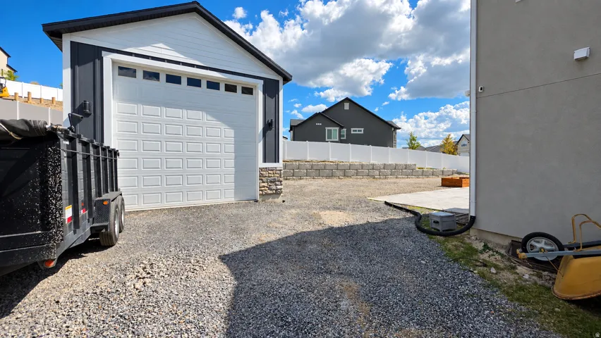 Detached garage with stone and vertical siding accents, white panel garage door with transom windows, gravel driveway, and a concrete patio area