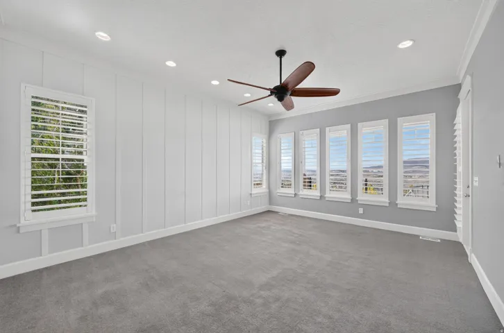 Recessed ceiling lighting, ceiling fan, multiple windows with plantation shutters, board and batten accent wall, and gray carpeting