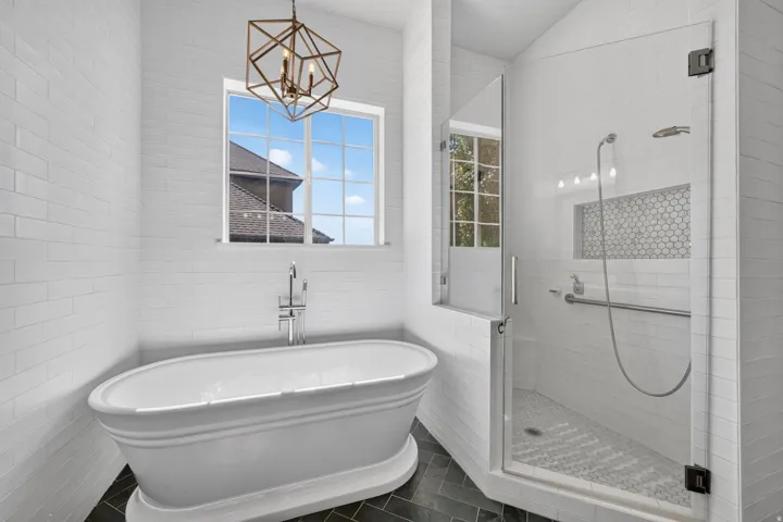 Freestanding soaking tub with chrome fixtures, glass-enclosed shower with a hexagon tile niche, geometric chandelier, and white rectangular wall tiling