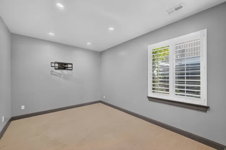 Recessed ceiling lighting, neutral wall paint, carpet flooring, a window with plantation shutters, and a wall-mounted television bracket