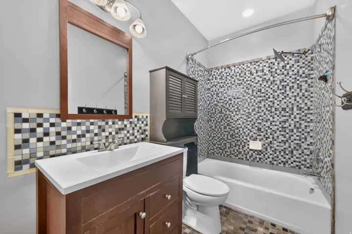 White vanity with a single basin sink and brushed nickel faucet, wood-framed mirror, mosaic tile backsplash, and a tiled shower with a curved rod