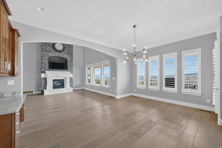Stone-faced fireplace with white mantel, wide plank wood-finish flooring, multiple windows with plantation shutters, vaulted ceilings, and a contemporary chandelier