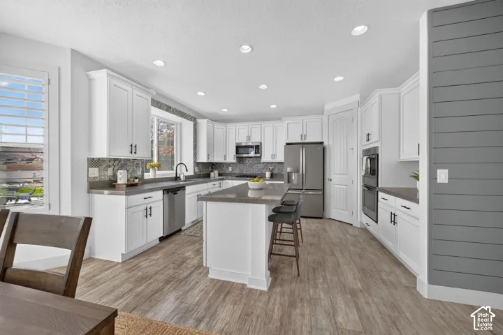 Kitchen featuring white cabinetry, stainless steel appliances, a central island with bar seating, wood-finish flooring, and a decorative shiplap accent wall